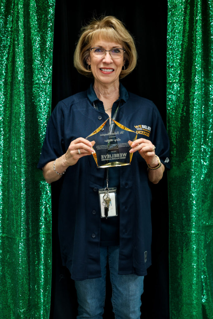 We Believe Champion holding a commemorative plaque in front of a green curtain
