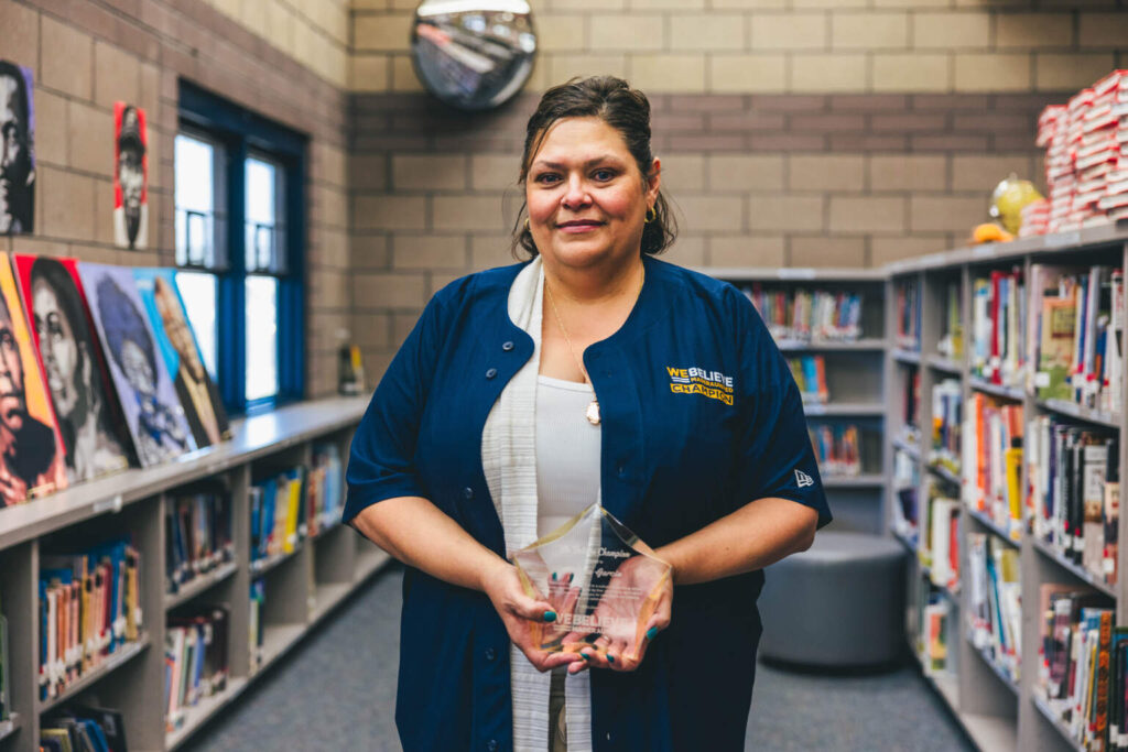 We Believe Champion holding a commemorative plaque in a school library