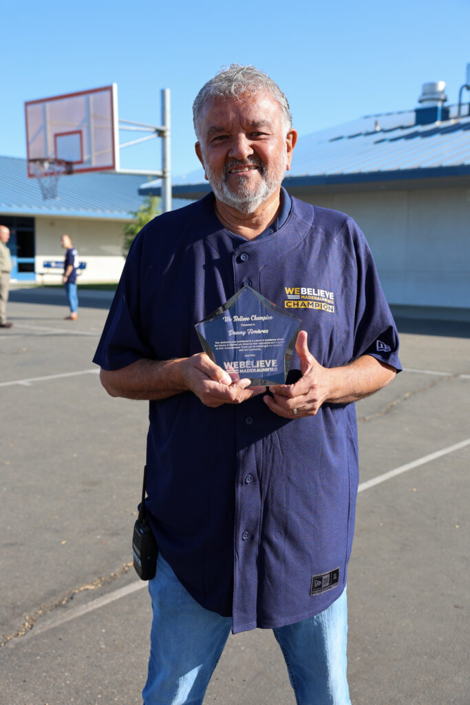 We Believe Champion holding a commemorative plaque on a school blacktop
