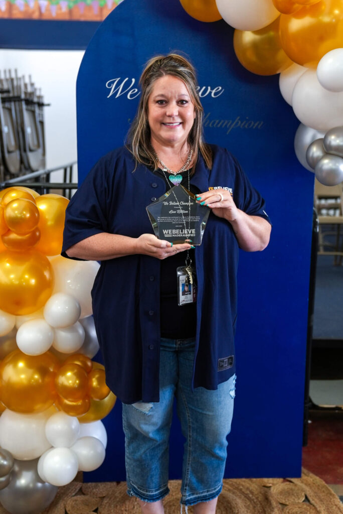 Lisa Killion, We Believe Champion, holding her commemorative plaque beside gold and white balloons
