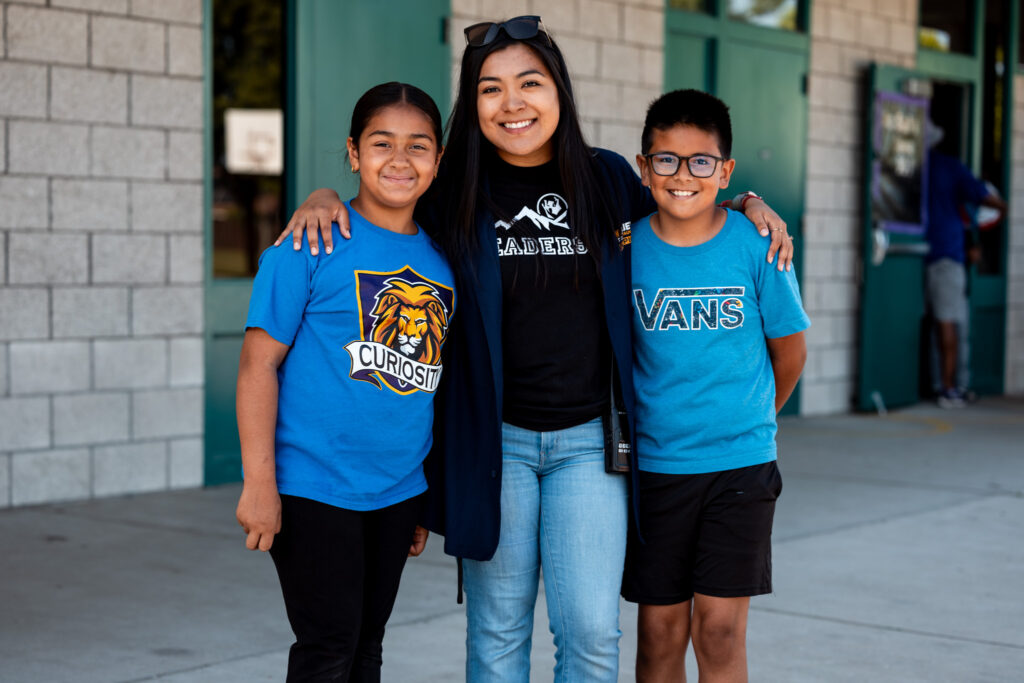 Maria Hernandez, We Believe Champion, smiling with two students outside a school building